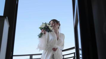 Bride holding a bouquet on a balcony with a blue sky, bride in a satin robe, holding a bouquet and smiling on a balcony. video