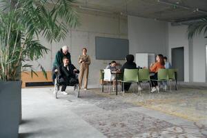 A diverse and multicultural business team collaborates in the office while their colleagues, including a director in a wheelchair and his friend, share a lighthearted moment, fostering an atmosphere photo