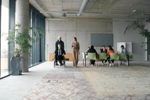 A diverse and multicultural business team collaborates in the office while their colleagues, including a director in a wheelchair and his friend, share a lighthearted moment, fostering an atmosphere photo