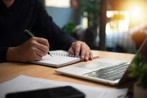 A man is writing on a notebook while sitting at a desk with a laptop and a pen photo