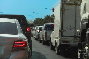 Rear sid of silver car with turn on brake light. Traffic of car line up in a queue during time of business or travel trip. Background of green tree and traffic light under blue sky. photo