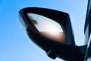 Under view of mirror wing on dark car with orange reflection in the mirror. Under clear blue sky. photo
