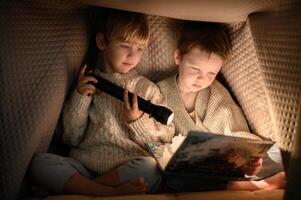 Two little boys whispering and reading a book under a blanket with a flashlight at night. photo
