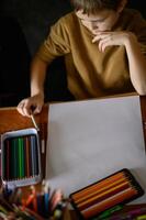 A boy sits at a table with colored pencils and markers, thinking about what to draw. photo