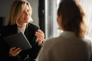 Two women in business attire having a conversation during a meeting. They are surrounded by laptops, documents, and a calm, professional office atmosphere. photo
