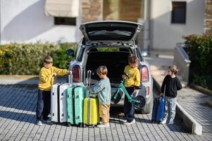 Four children stand near a packed car trunk, ready for an adventure with their bright luggage and scooters photo
