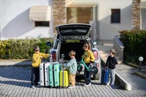 Four children stand by the cars open trunk surrounded by colorful suitcases and scooters ready for a trip photo