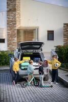The car trunk is open as four kids gather around, packing bright suitcases and their scooters for the journey. photo