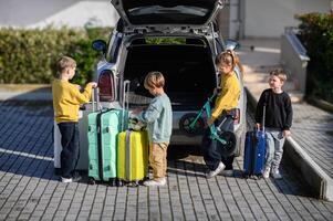 The car is parked with the trunk open as four kids gather around colorful suitcases and scooters, excited for their trip photo