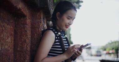 Attractive woman leaning against a wall on a sidewalk enjoying the city view while using a map app to find directions on mobile phone. video