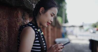 Handheld shot, Asian woman leaning against a wall on a sidewalk enjoying the city view while using a map app to find directions on mobile phone. video
