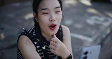 Handheld high angle view shot, Cheerful Asian woman sits on an outdoor sidewalk and enjoy a fun call with her friends via smartphone. video