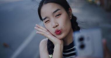 Handheld high angle view shot, Happy Asian woman posing for a selfie on her smartphone while relaxing on an outdoor sidewalk. video