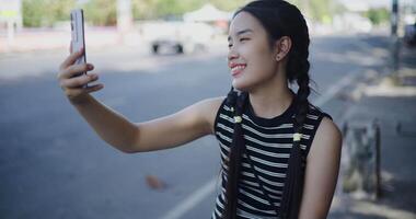 Handheld shot. Happy Asian woman posing for a selfie on her smartphone while relaxing on an outdoor sidewalk. video