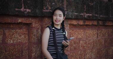 Attractive Asian woman leaning against a wall on a sidewalk enjoying the city view while using a map app to find directions on mobile phone. video