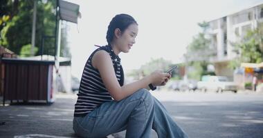 Handheld low angle view shot, Cheerful Asian woman sits on the sidewalk enjoying the city view and looking at something on her mobile phone. video