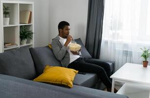 African American man sits alone on the couch and enjoying popcorn while watching television and streaming service photo