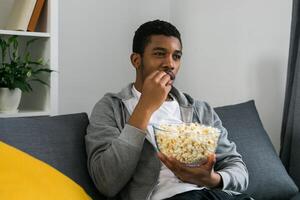 African American man sits alone on the couch and enjoying popcorn while watching television and streaming service photo