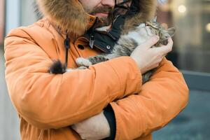 Bearded man in a fur lined hood smiles while holding a fluffy tabby cat outdoors close-up. Cozy and joyful winter scene in an urban environment, homeless animals photo