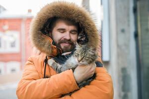 Bearded man in a fur lined hood smiles while holding a fluffy tabby cat outdoors. Cozy and joyful winter scene in an urban environment, homeless animals photo