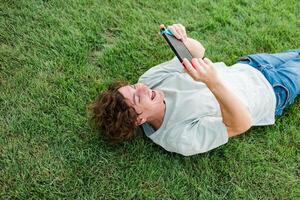 Joyful man smiling and having fun while playing games with portable console and lying on the grass outdoors. Technology and game addiction concept. photo
