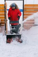 Red snow blower in action. Man outdoor in front of house using snowblower machine for removing snow on yard. Snow thrower in winter outside home. Young worker guy blowing snow photo