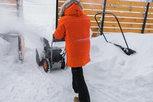Red snow blower in action. Man outdoor in front of house using snowblower machine for removing snow on yard. Snow thrower in winter outside home. Young worker guy blowing snow photo