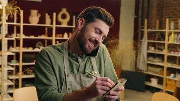 Busy smiling Caucasian man male entrepreneur potter happy small business owner artist businessman guy in apron talk mobile phone call client order writing notes pencil inside pottery ceramic workshop video