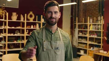 Happy smiling Caucasian small business owner man male guy joyful businessman salesman potter looking at camera showing thumb up gesture approval recommend reaction nice good indoors pottery workshop video