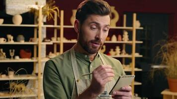 Pensive Caucasian salesman seller potter male man guy businessman artist small business owner in apron writing notes pencil think planning sell list idea notebook insight inside workshop store pottery video