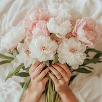 Soft pink and white peonies held gently in hands on a cozy bed photo