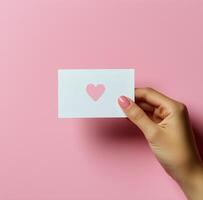 Hand holding a card with heart shape cutout against a pink background during creative display photo