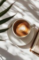 Warm cup of coffee placed beside an open book on a sunny table with shadows photo