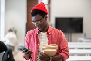 Satisfied hipster black student guy stands in college library with textbooks using phone break study photo