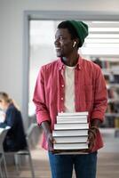 Cheerful positive African American man with stack of books walks through library enjoying studying photo