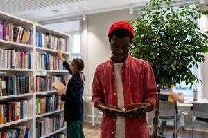 Smiling student black guy stands in library read book in background girl reach for top of bookshelf. photo