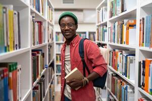 Smiling african american man student holds book in university library between bookshelves. photo