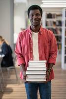 Portrait of happy African American student guy with pile of books in library photo