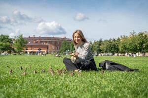 Smiling woman taking break in park, seated on grass, feeding sparrows with waffle cone ice cream photo
