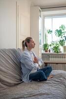 Woman sits on couch with eyes closed in lotus pose doing meditation practices, breathing exercises. photo