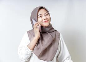 A smiling asian woman in a hijab talking on her phone, showcasing a cheerful demeanor isolated on white background photo