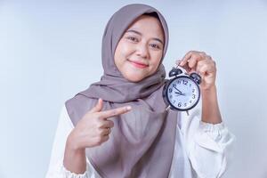 A smiling asian woman in a hijab holds an alarm clock, emphasizing the importance of time management, isolated on white background photo