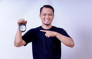 An asian man holding an alarm clock, joyfully pointing at it with a big smile isolated on white background photo