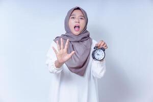 A surprised asian muslim woman holds an alarm clock, signaling a message about time urgency, isolated on white background photo