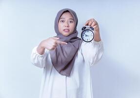 An asian woman in a hijab is pointing at a clock, expressing urgency or concern about time, isolated on white background photo