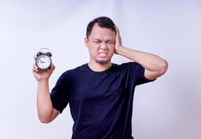 An asian man looks frustrated holding an alarm clock, feeling distressed by time pressure isolated on white background photo