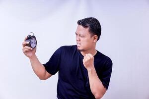 A frustrated asian man checks an alarm clock, expressing urgency or annoyance at the time isolated on white background photo