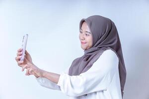 An asisan woman in a hijab takes a selfie, smiling happily isolated on white background photo