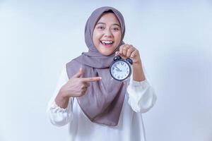 A cheerful asian woman in a hijab holds a clock and pointing to it, emphasizing the importance of time, isolated on white background photo