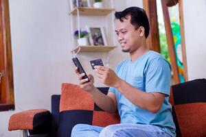 A young asian man is using his phone while holding a credit card, looking pleased with his purchase photo